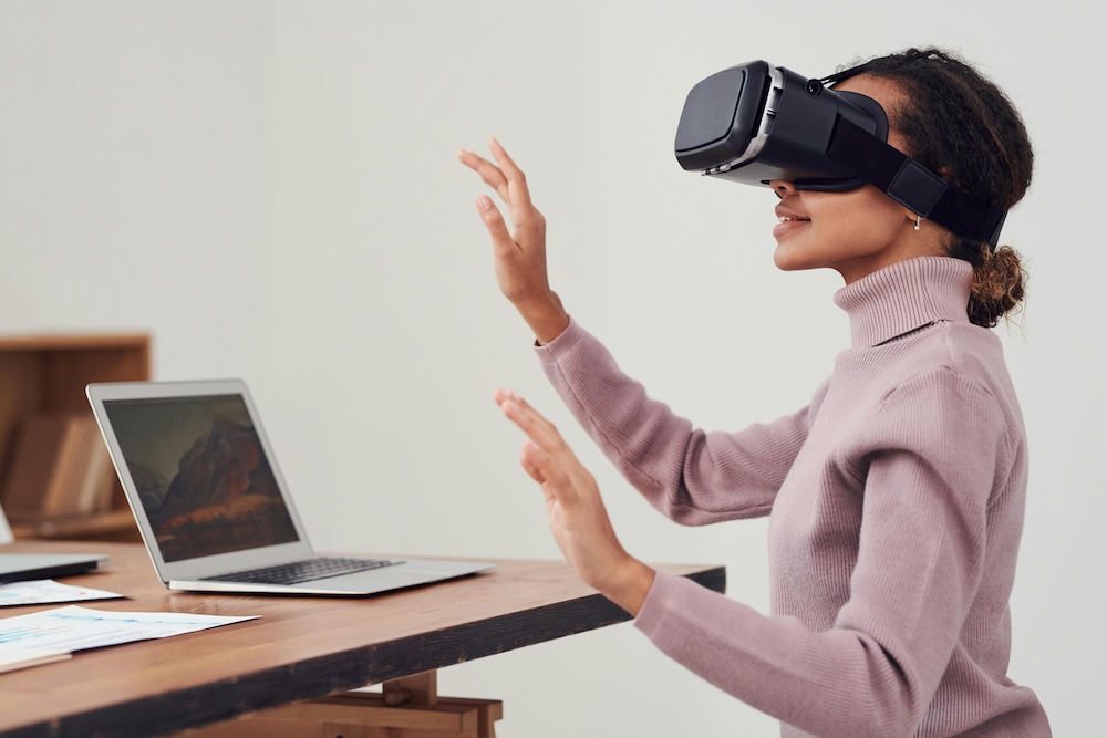 A woman wearing a virtual reality headset interacts with digital content while seated at a modern office desk with a laptop.